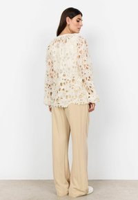 Krajkový top s dlouhým rukávem v off-white barvě s otevřenými květinovými vzory; doplněný o volné široké kalhoty béžové barvy a bílé obuví.