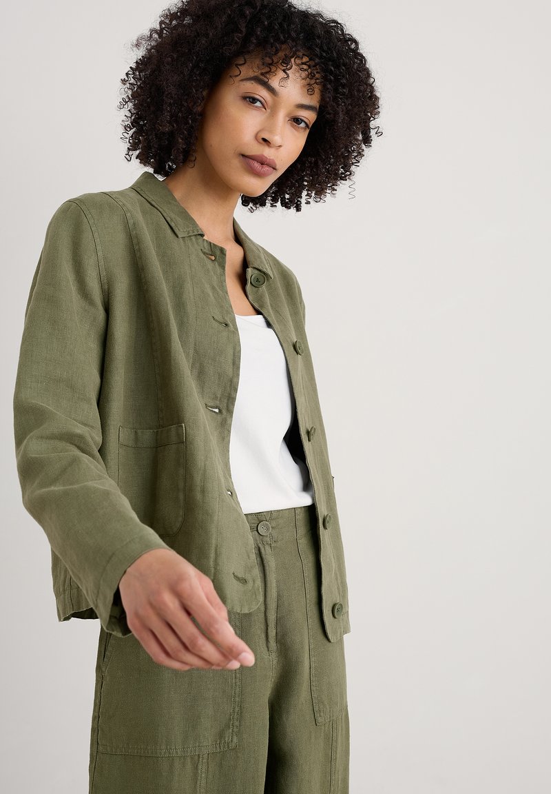 Chaqueta de lino verde con cuello, botones delanteros y bolsillos laterales, combinada con una camiseta blanca y pantalones de lino verdes a juego.