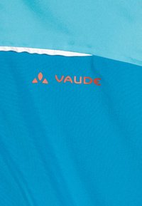 Turkizna tkanina z gladko površino, ki ima belo črto in oranžni logotip "Vaude". Oblikovanje je preprosto in moderno.