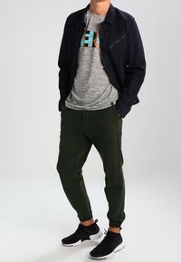 Czarna kurtka z kieszenią na zamek, szary t-shirt z kolorową grafiką, zielone zwężające się spodnie i czarne buty sportowe.