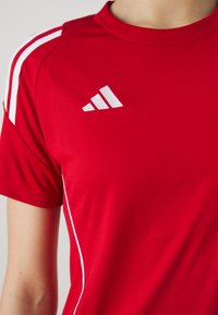 Camiseta atlética roja fabricada con una tela suave, con rayas blancas en los hombros y un logo bordado en blanco en el pecho.