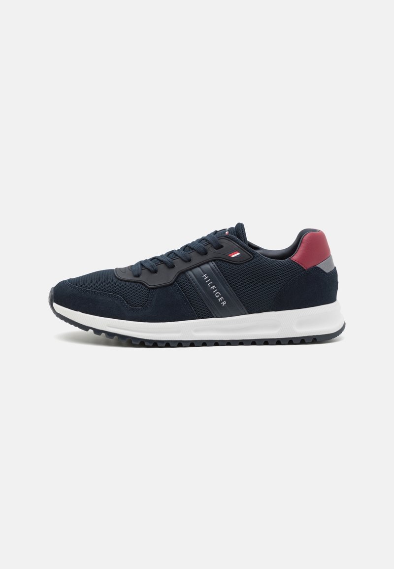 Baskets de sport bleu marine avec semelle blanche, accent rouge au talon, textures en mesh et suède, lacets, et marquage "Hilfiger" sur le côté.