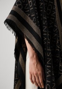 Zwart en gouden poncho met een patroon en franjes, gekenmerkt door gedurfde strepen en geweven tekstdetails, tegen een neutrale achtergrond.