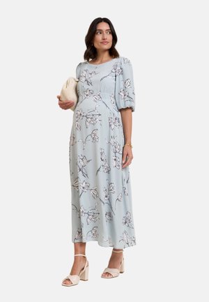 Femme enceinte portant une robe midi fleurie bleu clair avec des manches bouffantes, tenant une pochette beige texturée et portant des talons blocs beiges.