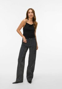 Crna bezrukavna majica s cowl izrezom, uparena s sjajnim crnim hlačama širokog kroja s točkastim uzorkom i cipelama s oštrim vrhom.