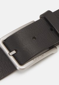 Cintura in pelle nera con fibbia in metallo argentato, caratterizzata da una finitura testurizzata e cinque regolazioni a foro. Logo Calvin Klein inciso sulla fibbia.