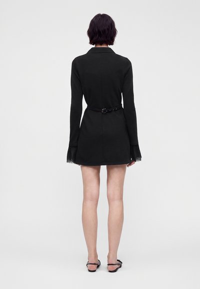 Femme aux cheveux courts et foncés portant une mini-robe noire à manches longues avec une ceinture à la taille et des poignets transparents, debout de dos sur un fond uni.