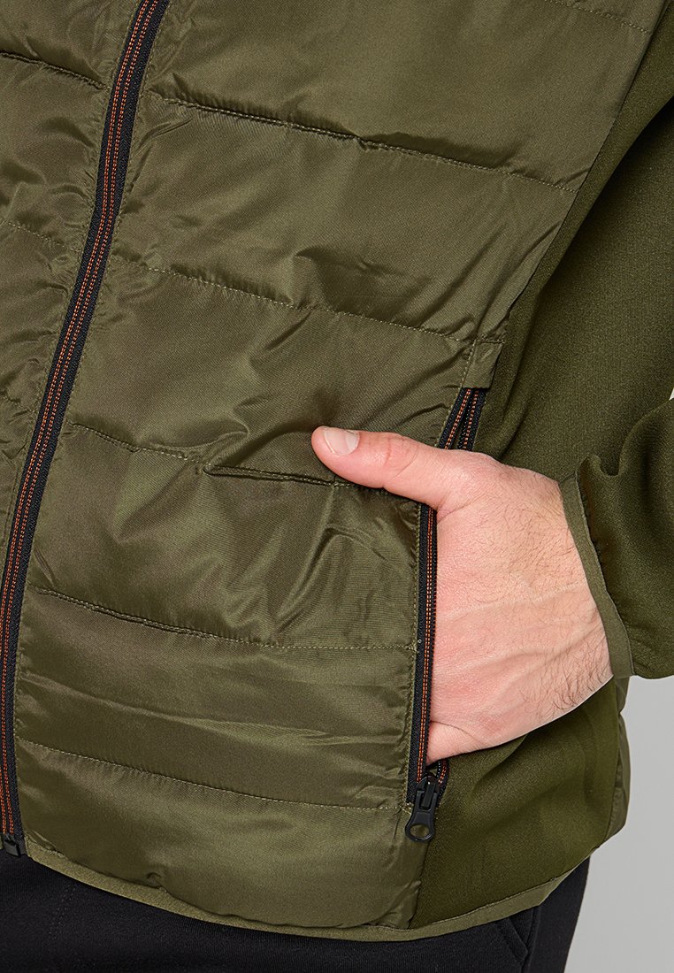 Ręka wsuwająca się do zapinanej na zamek kieszeni oliwiono-zielonej pikowanej kurtki z czarnymi i pomarańczowymi detalami zamka.