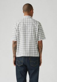 Camisa de manga corta en blanco con patrón de rombos grises, diseño de cuello y una tela ligera. La vista trasera muestra un corte holgado y un dobladillo liso.