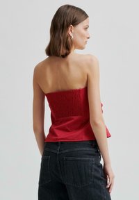 Czerwona bluzka bez ramiączek wykonana z gładkiego materiału, z marszczonym tyłem i rozkloszowanym dołem. W parze z ciemnoniebieskimi dżinsami z wysokim stanem.