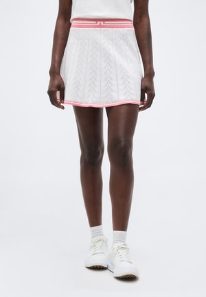 Γυναίκα που φοράει λευκή φούστα τένις με ροζ περίγραμμα, λευκές κάλτσες και λευκά αθλητικά παπούτσια, στέκεται απέναντι σε απλό φωτεινό φόντο.