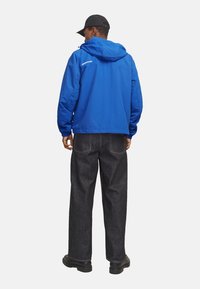 Blaue Kapuzenjacke mit elastischen Bündchen, kombiniert mit dunklen Jeans. Die Jacke verfügt über ein Logo auf dem Rücken und das Design ist leger geschnitten.