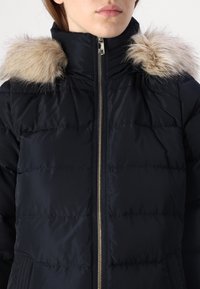 Giacca trapuntata blu navy con zip frontale, dotata di cappuccio rifinito in delicata pelliccia sintetica. Il tessuto è liscio e trapuntato, con tasche laterali.