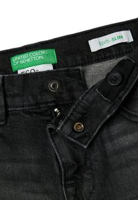 Jean en denim noir avec une coupe slim et une finition légèrement texturée. Comprend une fermeture à bouton et des étiquettes de marque, l'une en vert et l'autre en blanc.