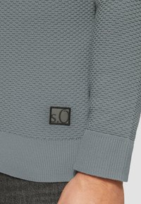 Hellgrauer Strickpullover mit strukturiertem Muster, gerippten Bündchen und einem schwarzen Logopatch an der Seite. Glattes Material und taillierte Passform.