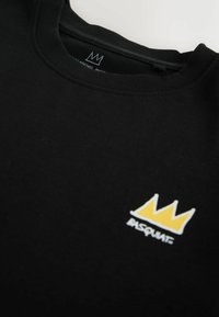 Fekete, kerek nyakú póló kis sárga koronás grafikával és az alatta, a mellrészen nyomtatott "BASQUIAT" felirattal.