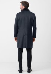 Donker wollen overjas met een met bont gevoerde kraag, op maat ontworpen, rechte snit en zij vents, gecombineerd met zwarte slim-fit broek en leren laarzen.