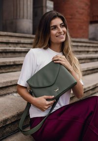 Zelená kožená kabelka s klopovým designem, zlatými kovovými detaily a ramenním popruhem. Modelka má na sobě bílou košili a vínovou sukni.