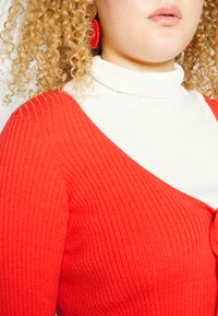 Sweater vermelho canelado com decote em V, sobre um suéter branco canelado de gola alta. Cabelo encaracolado e brincos redondos vermelhos são visíveis.