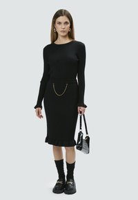Vestido de punto negro con mangas largas, dobladillo con volantes y detalle de cadena dorada en la cintura. Combinado con zapatos de plataforma negros y un bolso con estampado de cocodrilo.