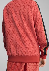Rot gemusterte Jacke mit geometrischen Designs, gerippten Bündchen und einem einfarbigen marineblauen Seitenstreifen mit gelbem Akzent entlang der Ärmel.