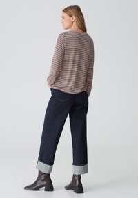 Langärmliges Oberteil mit einem geometrischen Muster in Rot und Creme, kombiniert mit dunkelblauen, weit geschnittenen Jeans, die an den Knöcheln umgeschlagen sind, und braunen Stiefeletten.