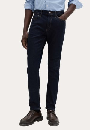 Mężczyzna ubrany w ciemnoniebieskie dopasowane dżinsy, jasnoniebieską podwiniętą koszulę i ciemnobrązowe skórzane buty, stojący z jedną ręką w kieszeni.