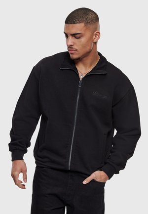 Homme aux cheveux courts portant une veste noire zippée avec le logo "Dropize", une main dans la poche et regardant vers le bas sur le côté.