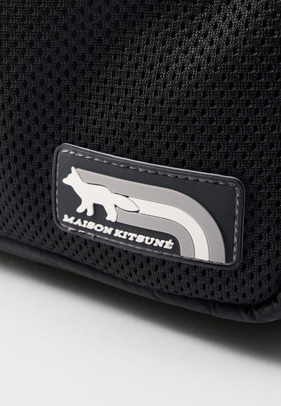 Sac en maille noir avec un patch rectangulaire représentant un logo de renard blanc et le texte "MAISON KITSUNÉ", sur fond de surface texturée.