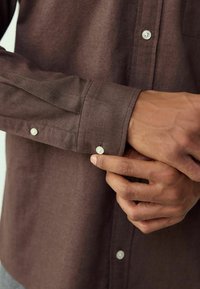 Mörkbrun långärmad skjorta med en slät textur. Har vita knappar vid manschetterna och knappraden. Närbild på hand som justerar manschetten.