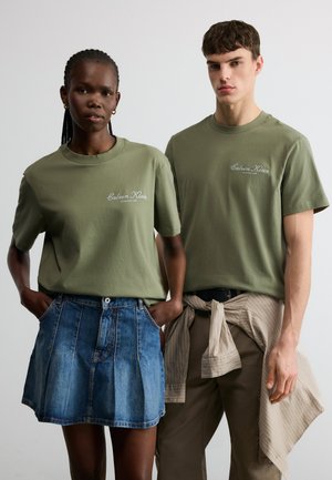 Doi tineri adulți purtând tricouri Calvin Klein verde măsliniu; unul într-o fustă albastră din denim, celălalt cu o cămașă bej legată în jurul taliei.