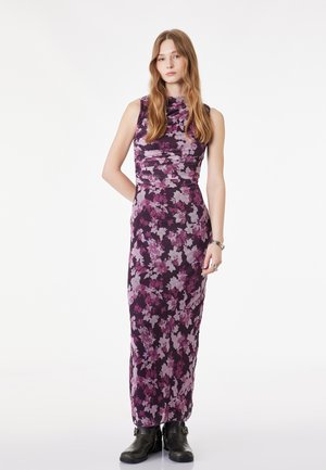 Junge Frau mit langen Haaren, die ein ärmelloses lila Maxikleid mit Blumenmuster und schwarze Stiefel trägt, steht vor einem weißen Hintergrund.