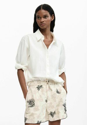 Donna con lunghi capelli scuri indossa una camicia bianca ampia con bottoni e maniche arrotolate e pantaloncini beige con motivi floreali neri e chiari.