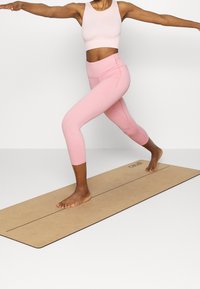 Rosa sport-bh och capri-leggings i stretchtyg, framförd på en beige yogamatta med branding, barfota, i en krigarställning.