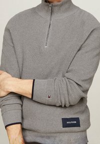 Grauer Rippstrickpullover mit Viertelreißverschluss, kleinem Flaggenlogo am Ärmelbündchen und "Hilfiger"-Label nahe dem Saum, getragen von einer Person mit verschränkten Armen.
