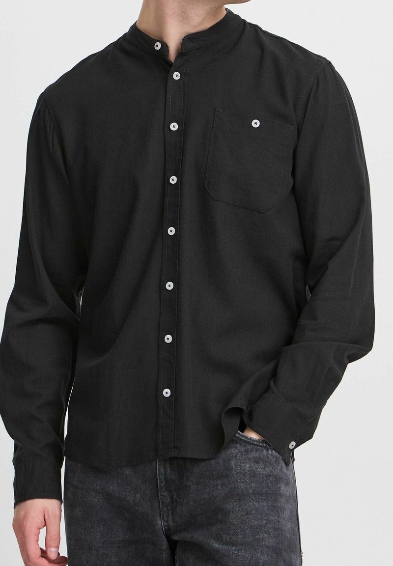 Homme portant une chemise noire à col mandarin avec des boutons blancs et une poche poitrine, associée à un jean gris foncé, main dans la poche.
