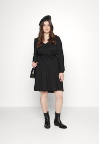 Vestido preto de manga longa com decote em V e detalhe de cintura amarrada, combinado com botas de tornozelo pretas e um boina preta. Tecido suave, até ao joelho.