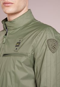 Veste légère vert olive avec un col montant, une poche poitrine zippée et un patch brodé sur l'épaule. Tissu lisse et texturé.