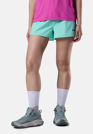 Persoon die mintgroene korte broek, een roze top, witte sokken tot halverwege de kuit en turquoise wandelschoenen draagt, staand tegen een effen achtergrond.