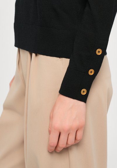Sort striktrøje med lige kant og tre træknapper på ærmet, parret med beige bukser. Glat tekstur og tætsiddende design.