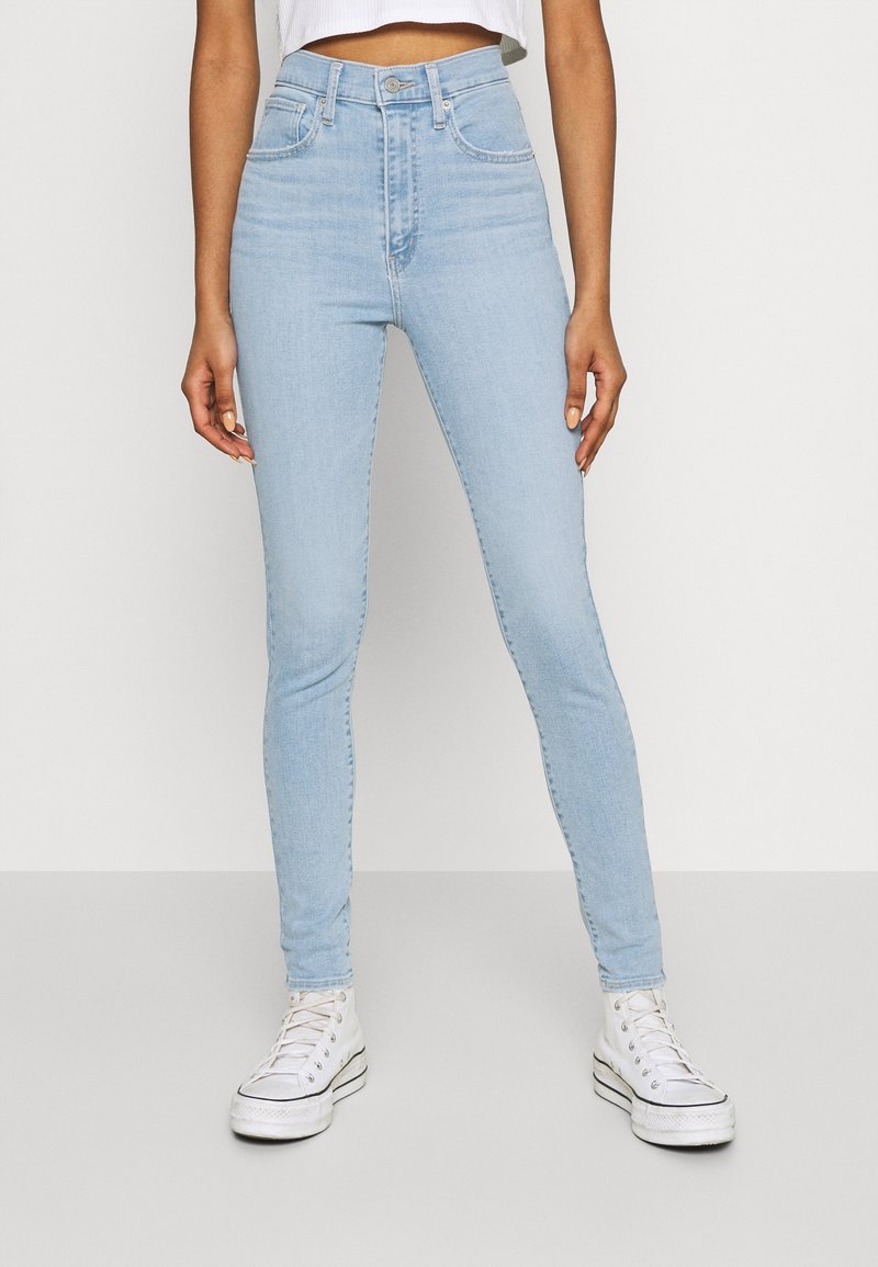 Personne portant un jean skinny taille haute bleu clair et des baskets blanches montantes, debout sur un fond gris uni.