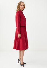 Veste taillée rouge avec poches avant et boutons, assortie à une robe évasée. Le mannequin porte des escarpins noirs à bout pointu.