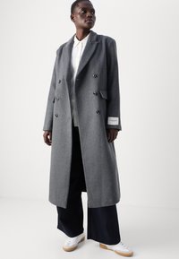 Caban lung din lână gri, cu un design dublu, două buzunare frontale și nasturi negri. Purta peste o cămașă albă și pantaloni negri largi.