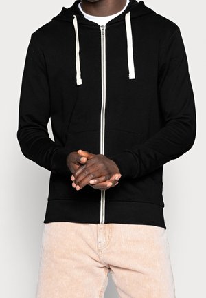 Hombre vestido con una sudadera con cremallera negra y cordones blancos, y pantalones de pana beige, con las manos entrelazadas a la cintura.