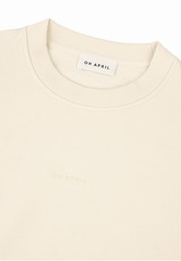 Sweat-shirt de couleur crème avec un col côtelé, présente un logo discret en relief "OH APRIL" près de l'ourlet, fabriqué en matériau doux.