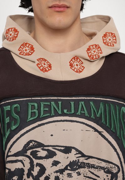 Sweat-shirt à capuche avec une capuche beige ornée de motifs brodés orange. Le corps principal est marron foncé avec un texte vert et une impression graphique noire.