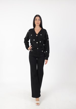 Cardigan noir avec des motifs de cœurs blancs, décolleté en V et fermeture à boutons dorés, associé à un pantalon noir taille haute. La modèle se tient devant un fond neutre.