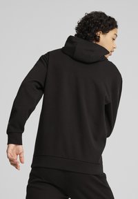 Sudadera negra con capucha, de manga larga, textura suave y corte holgado. La parte trasera presenta un diseño sin costuras y un sutil logotipo cerca del dobladillo.
