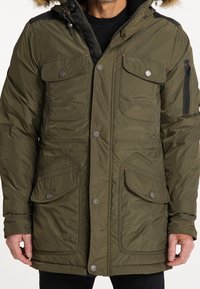 Oliwkowo-zielona ocieplana parka z zamkiem błyskawicznym i zapięciem na zatrzaski, licznymi kieszeniami z przodu, kapturem podszytym futrem oraz kontrastującymi czarnymi akcentami na ramionach.