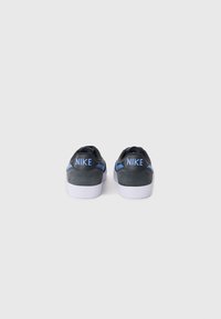 Baskets basses en daim gris avec une semelle en caoutchouc blanche, arborant un logo Nike bleu à l'arrière et une bande d'accent bleu sur les côtés.
